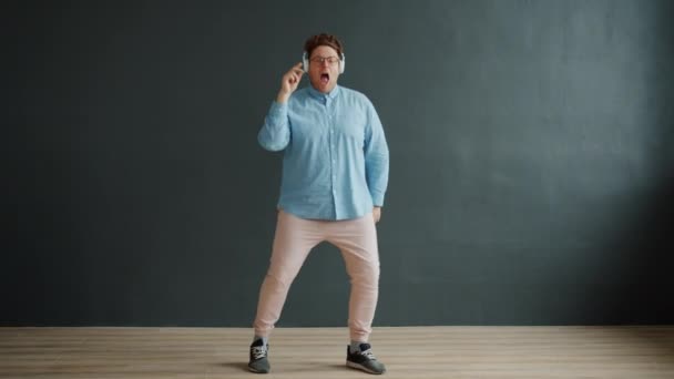 Hombre atractivo bailando en interiores divirtiéndose usando auriculares sobre fondo oscuro — Vídeos de Stock