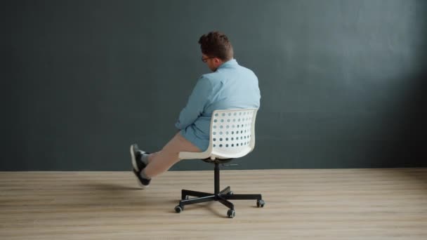 Грустный молодой человек, крутящийся в офисном кресле в помещении студии на темном фоне — стоковое видео