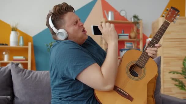 Slow motion of man playing guitar singing in smartphone wearing headphones at home — 비디오