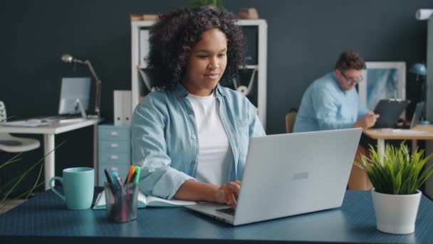 Joven dama afroamericana usando el ordenador portátil en la oficina, colega trabajando en segundo plano — Vídeo de stock