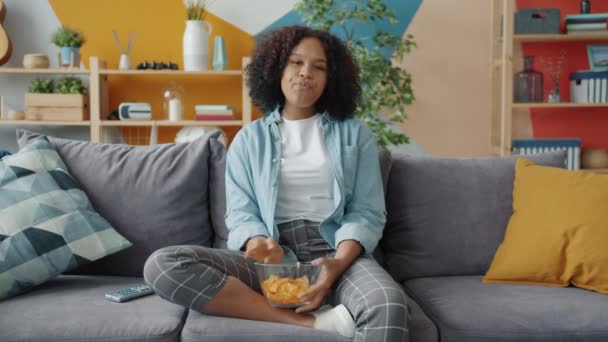 Retrato de una chica afroamericana aterrorizada viendo películas de terror en la televisión comiendo bocadillos — Vídeo de stock