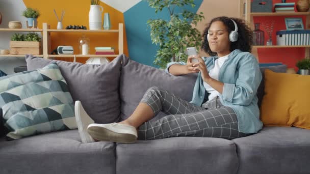 Alegre dama afroamericana usando teléfono inteligente disfrutando de la música en los auriculares en casa — Vídeo de stock