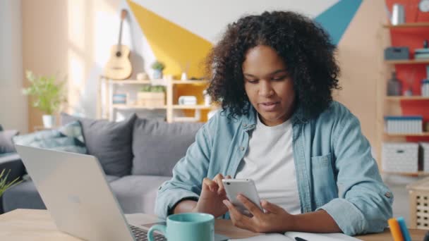 Hermosa dama afroamericana usando la pantalla táctil del teléfono inteligente sonriendo en casa — Vídeo de stock