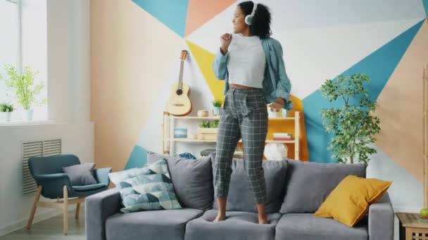 Chica afroamericana en audífonos bailando en sofá escuchando música divirtiéndose — Vídeos de Stock