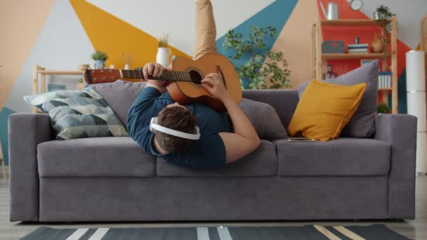 Chico alegre tocando la guitarra cantando usando auriculares divirtiéndose en el sofá en casa — Vídeo de stock