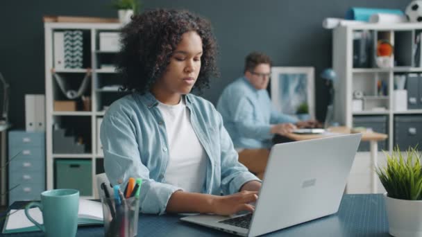 Menina afro-americana bonita trabalhando com laptop no escritório, em seguida, falando com colega — Vídeo de Stock