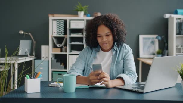 Hermosa dama afroamericana usando la pantalla táctil del teléfono inteligente sonriendo sentado en el escritorio — Vídeo de stock