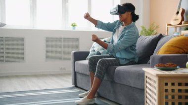 VR gözlüklü Afro-Amerikalı kadın evde kol ve bacaklarını oynatarak araba sürmekten zevk alıyor.