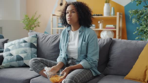 Emocjonalne mieszane rasy pani płacze oglądając telewizję w domu na kanapie gospodarstwa przekąski — Wideo stockowe