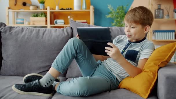 Veselý malý chlapec drží tablet pomocí moderního zařízení na gauči doma sám — Stock video