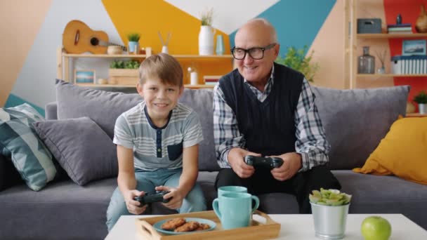 Abuelo y nieto están jugando video juego sentado en el sofá en casa — Vídeos de Stock