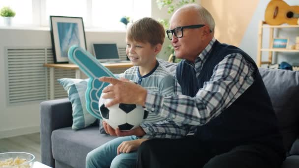 Заботливый дедушка и счастливый мальчик смотрят футбол по телевизору, держа в руках пену и мяч — стоковое видео