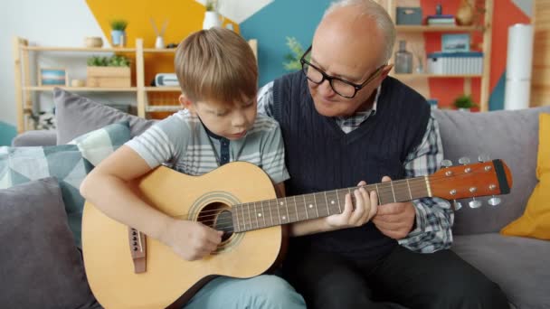 Old man teaching talented little grandson to play the guitar in apartment — Stock Video