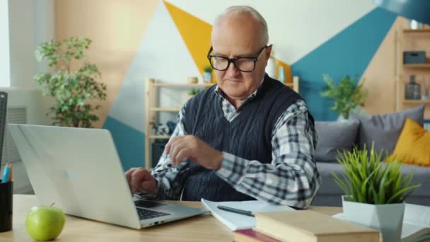 Oudere man met behulp van laptop computer typen gaan online het maken van aantekeningen thuis — Stockvideo