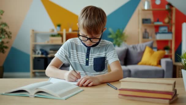 Retrato de niño alegre haciendo la tarea y luego mirando a la cámara sonriendo en casa — Vídeo de stock