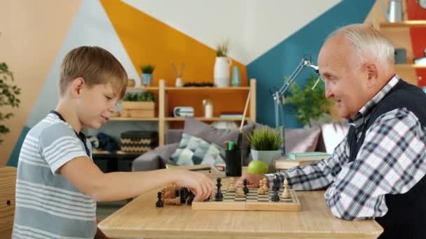 Hombre mayor y nieto divertido jugando ajedrez en casa haciendo risa choca esos cinco — Vídeo de stock