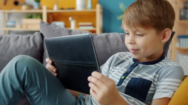 Cámara lenta del niño sonriente sosteniendo la tableta viendo video en el interior del apartamento — Vídeos de Stock