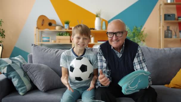 Football fans old man and child are watching match on TV at home holding ball — Stock Video