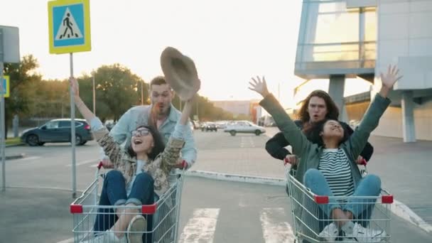 Uomini e donne che cavalcano carrelli della spesa nel parcheggio in città divertendosi a ridere — Video Stock