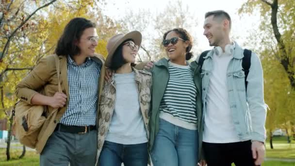 Молодые люди мультиэтничной группы прогуливаясь в парке с рюкзаками говорить смеясь — стоковое видео