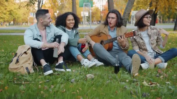 Carefree students singing playing the guitar relaxing on lawn in city park — 비디오