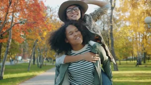 Lassú mozgás boldog lány így piggyback lovagolni örömteli hölgy barátja a parkban — Stock videók