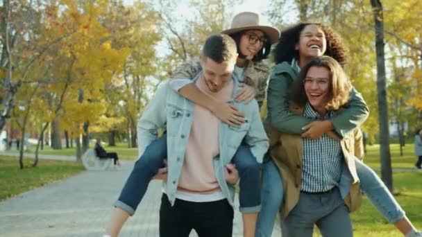 Jóvenes alegres dando paseo a cuestas a mujeres felices divirtiéndose en el parque — Vídeos de Stock