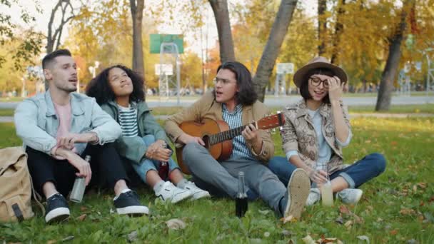 Retrato de chicas y chicos alegres cantando la guitarra en el césped en el parque de la ciudad — Vídeos de Stock