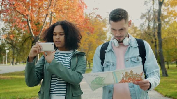 Турист-мужчина изучает карту, в то время как афроамериканка пользуется смартфоном на открытом воздухе — стоковое видео
