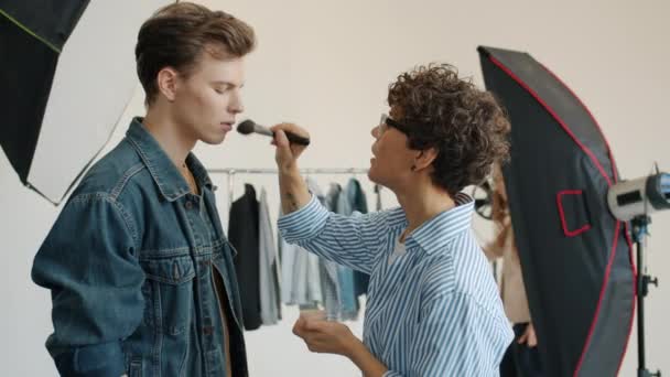 Mujer maquilladora ocupada con modelo masculino embellecer chico en el interior en el estudio — Vídeo de stock