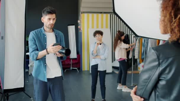 Fotógrafo sério tirando fotos de modelo de raça mista em estúdio falando usando câmera — Vídeo de Stock