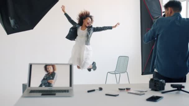 Счастливая молодая женщина прыгает весело позируя перед камерой в студии с фотографом — стоковое видео