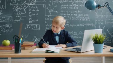 Smart little boy writing at desk in class, formulas are on chalkboard in background