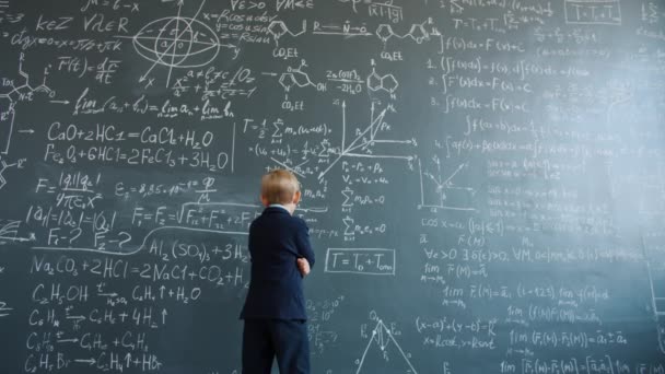 Little boy thinking looking at formulas written on chalkboard wall in college — Stockvideo