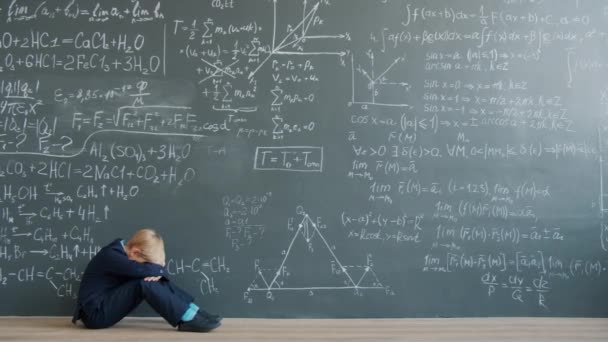 Sad child sitting on floor near chalkboard wall thinking solving problem alone — Stockvideo