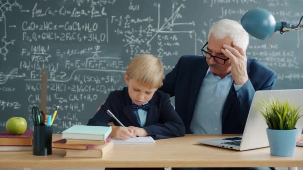 Mały chłopiec uczący się z nauczycielem piszącym przy biurku w klasie skupiony na edukacji — Wideo stockowe