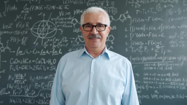 Elderly professor smiling looking at camera standing near blackboard with formulas — Stockvideo