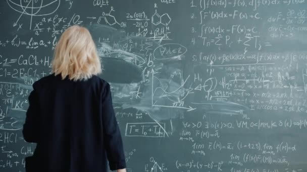 Back view of female scientist walking to chalkboard writing formulas with chalk — Stockvideo