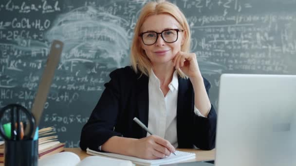 Amichevole signora matura sorridente seduta alla scrivania in classe guardando la fotocamera al chiuso — Video Stock