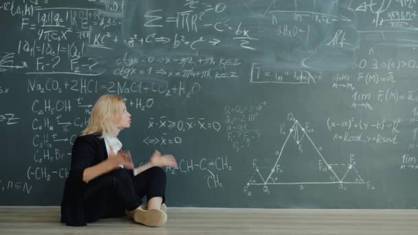 Langzame beweging van emotionele volwassen vrouw professor zitten op de vloer in de buurt van schoolbord — Stockvideo