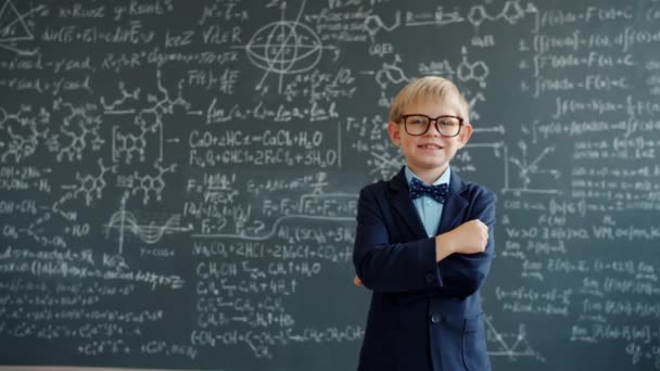 Boldog gyerek öltönyben és szemüvegben áll az osztályban táblával a háttérben — Stock videók
