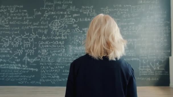 Female scientist walking to chalkboard wall with formulas in university classroom — Stockvideo