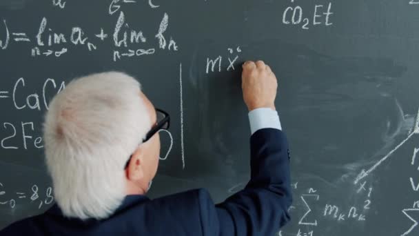 Vista posteriore di intelligente professore senior che scrive equazioni sulla lavagna in classe — Video Stock
