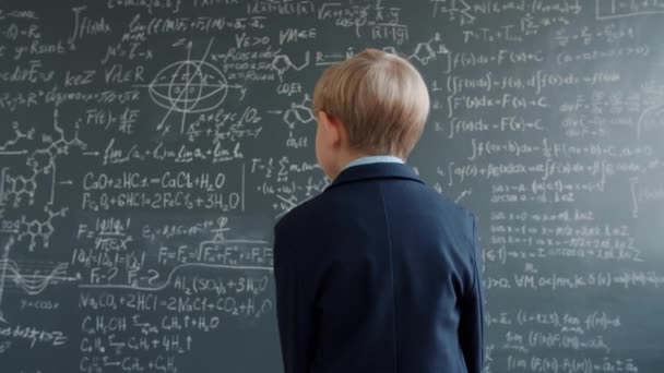 Dolly shot of little student walking to chalkboard looking at formula thinking — 图库视频影像