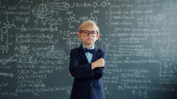 Préscolaire sérieux en costume et lunettes debout en classe près du tableau noir avec des formules — Video