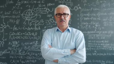 Serious scientist standing indoors with arms crossed near chalkboard with formulas
