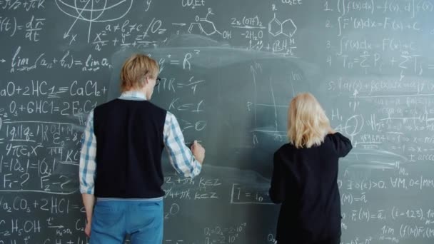 Man and woman writing formulas on chalkboard working at science project together — Stockvideo