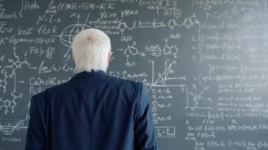 Mature man in suit walking to chalkboard wall with formulas looking at equations