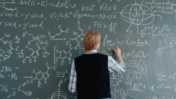 Back view of young maths teacher writing equations on chalkboard in classroom — 图库视频影像