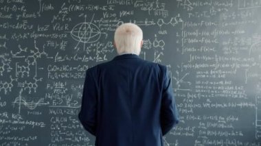 Experienced scientist walking to blackboard wall in college class looking at formulas